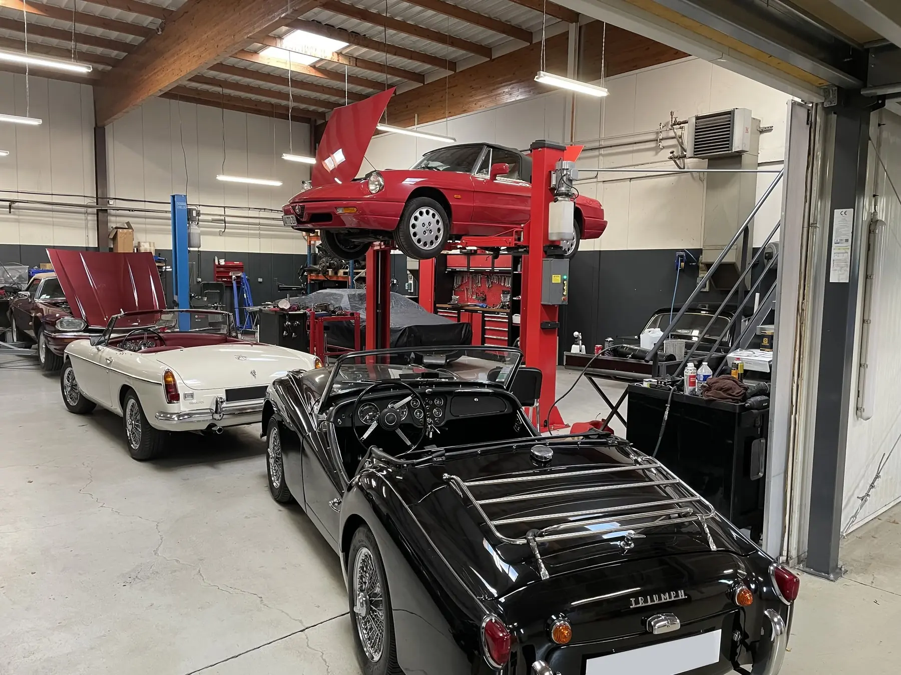 Alfa Romeo Spider sur pont élévateur, Triumph TR3 et MGB cabriolet à l'atelier mécanique